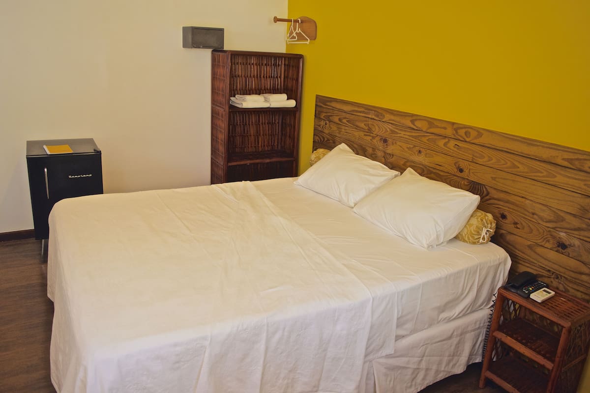 Een net opgemaakt bed met witte lakens, een houten hoofdbord, een klein nachtkastje met een telefoon en een houten garderobe met kledingroede.
