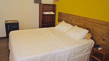 Cama bem arrumada com lençóis brancos, cabeceira de madeira, mesa de cabeceira pequena com um telefone e guarda-roupa de madeira com cabides.