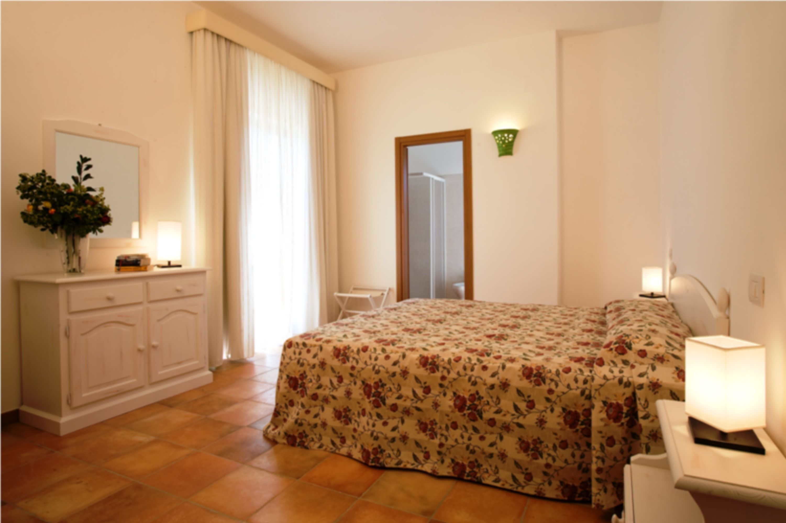 Une chambre à coucher avec une couvre-lit à motifs floraux, une commode avec un miroir, une table de chevet avec une lampe et une fenêtre avec des rideaux.