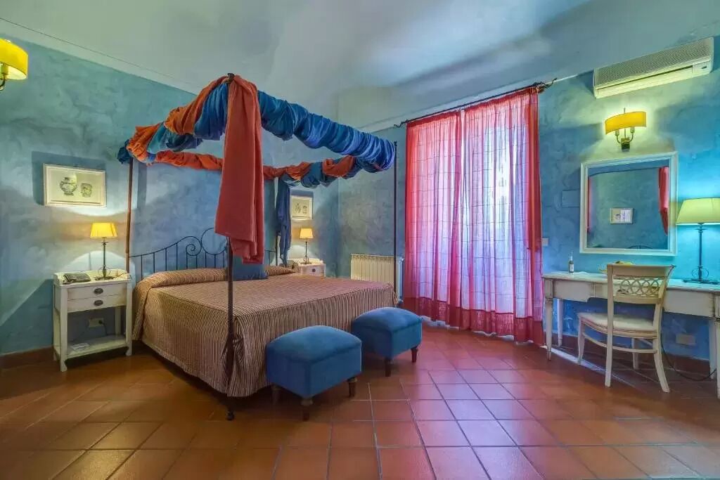 A bedroom with a four-poster bed, two blue ottomans, a desk with a chair, and a window with red curtains.