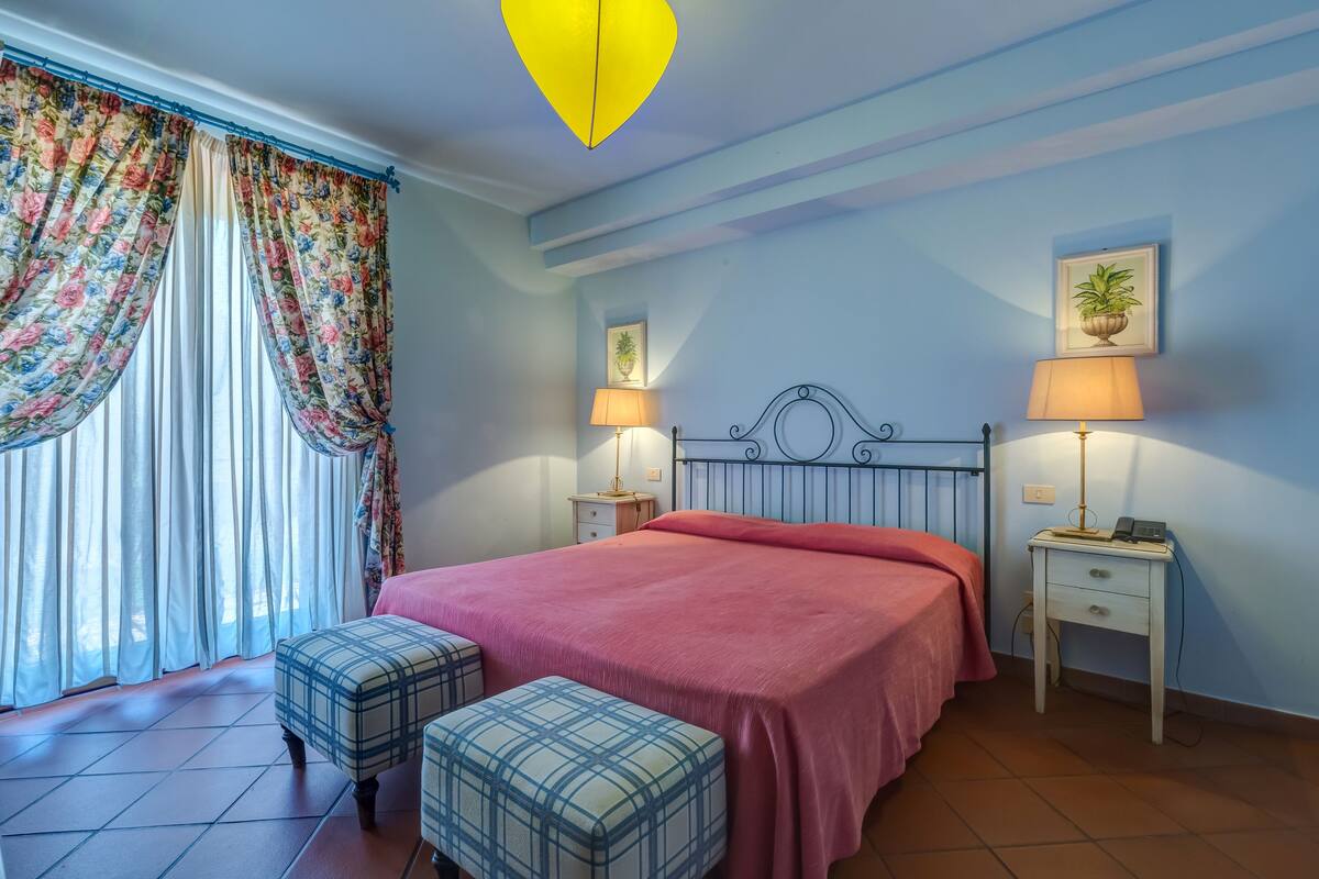 Une chambre à coucher avec un lit, deux tables de chevet avec des lampes, une fenêtre avec des rideaux et une petite plante en pot accrochée au mur.