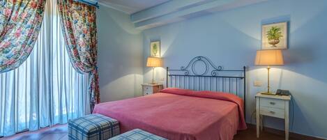 A bedroom with a bed, two bedside tables with lamps, a window with curtains, and a small potted plant on the wall.