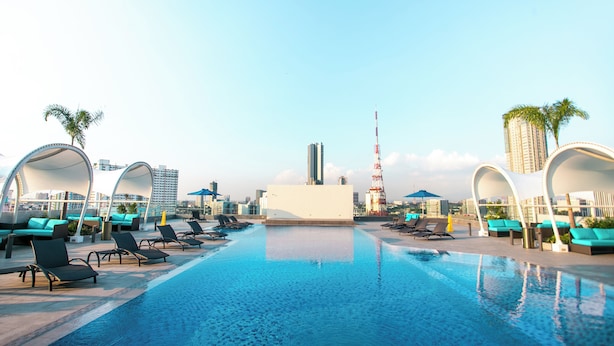 A rooftop pool with lounge chairs and umbrellas, surrounded by modern buildings and a tall tower. - Luxent Hotel (Quezon City)