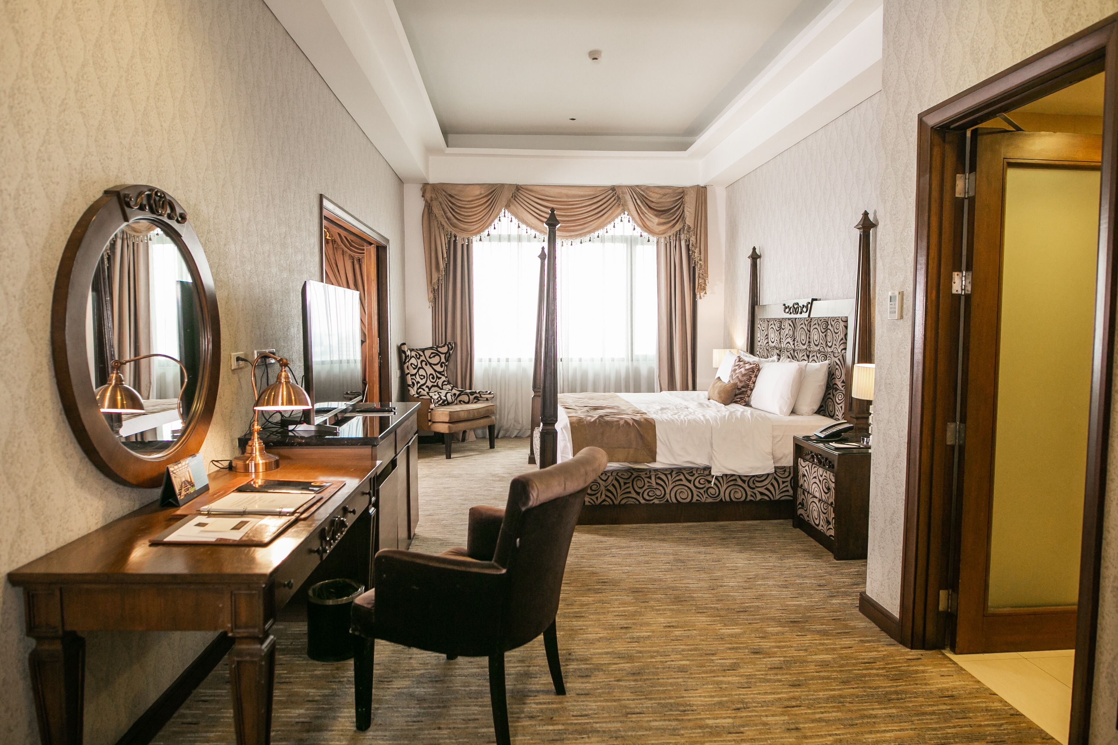 A hotel room with a large bed, a desk with a mirror, a chair, a nightstand, and a wardrobe.