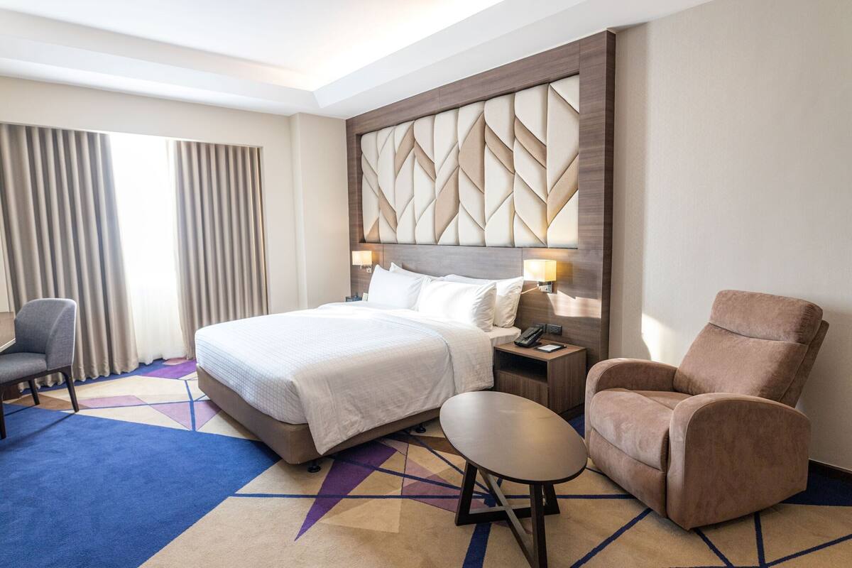 A hotel room with a large bed, a chair, a small table, and a nightstand with a lamp.