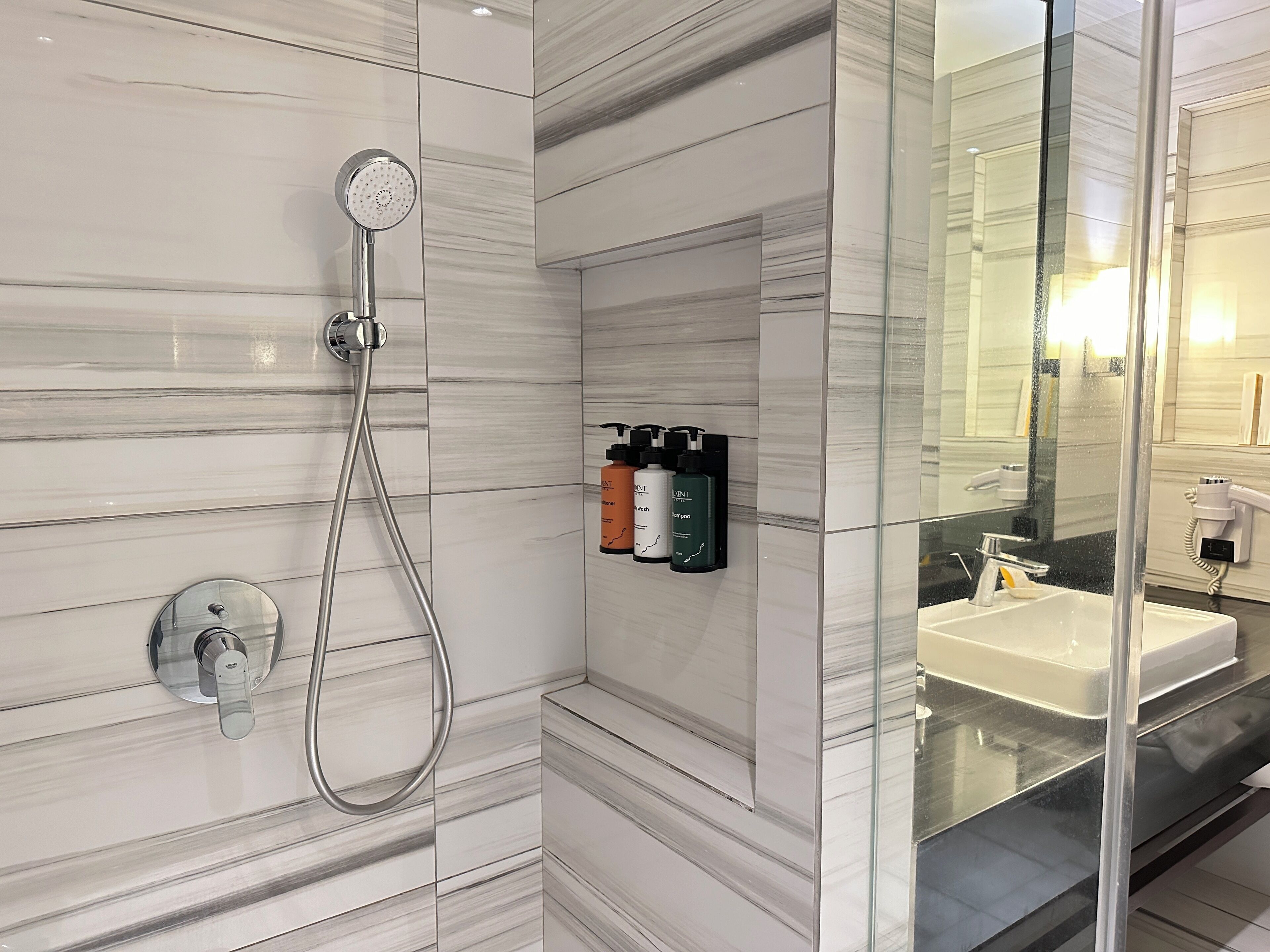 Un bagno moderno con box doccia in vetro, erogatore a muro per shampoo e balsamo e un lavandino ben visibile con specchio.