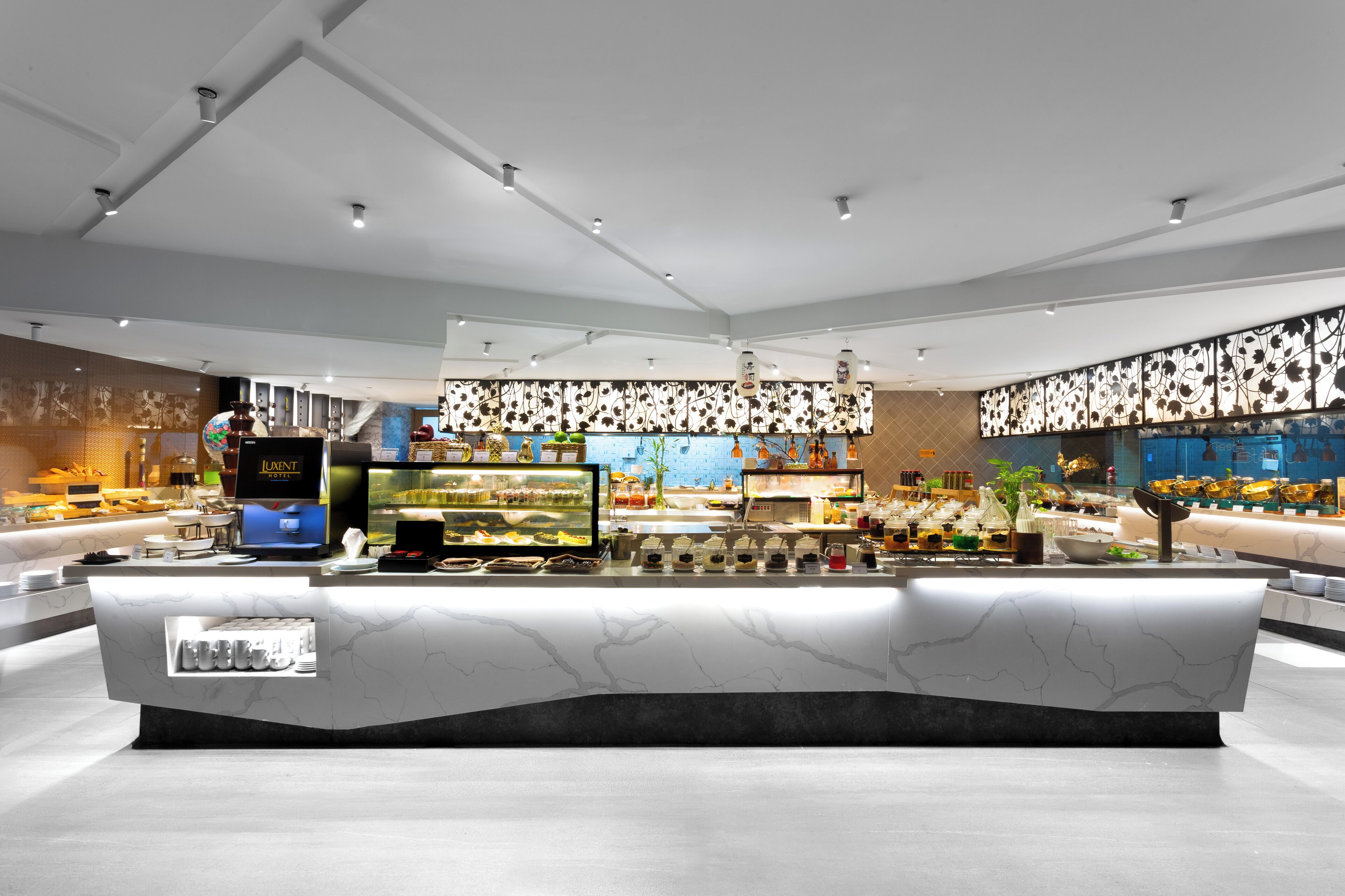 A modern hotel lobby with a marble reception desk, a coffee machine, and a variety of food displays.