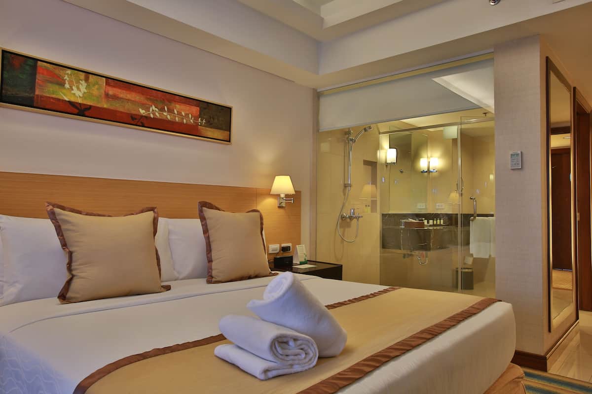 A hotel room with a large bed, a painting on the wall, a nightstand with a lamp, and a glass-enclosed shower area.