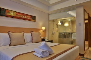 Habitación de hotel con una cama grande, un cuadro en la pared, una mesita de noche con lámpara y una zona de ducha con mampara de vidrio.