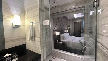 Un baño de hotel moderno con ducha de vidrio, tocador con encimera negra, y una cama visible a través de la puerta de la ducha.