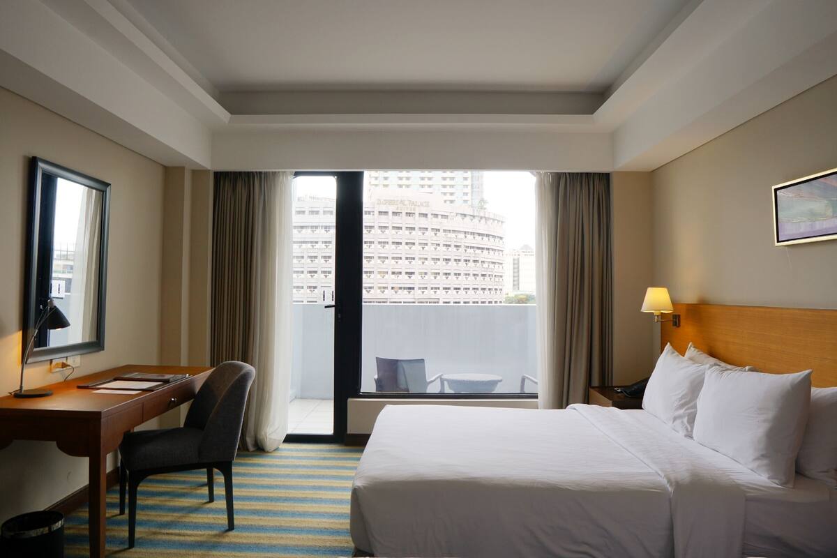 A hotel room with a bed, a desk, a chair, a lamp, and a view of a building through the window.