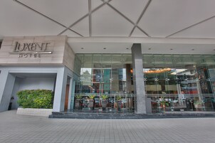 El exterior del Hotel Luxent con fachada de vidrio moderna, un área ajardinada con plantas y una vista despejada del interior a través del vidrio.