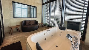 A modern bathroom with a large bathtub, a small sofa, and a window with blinds. - Luxent Hotel (Quezon City)