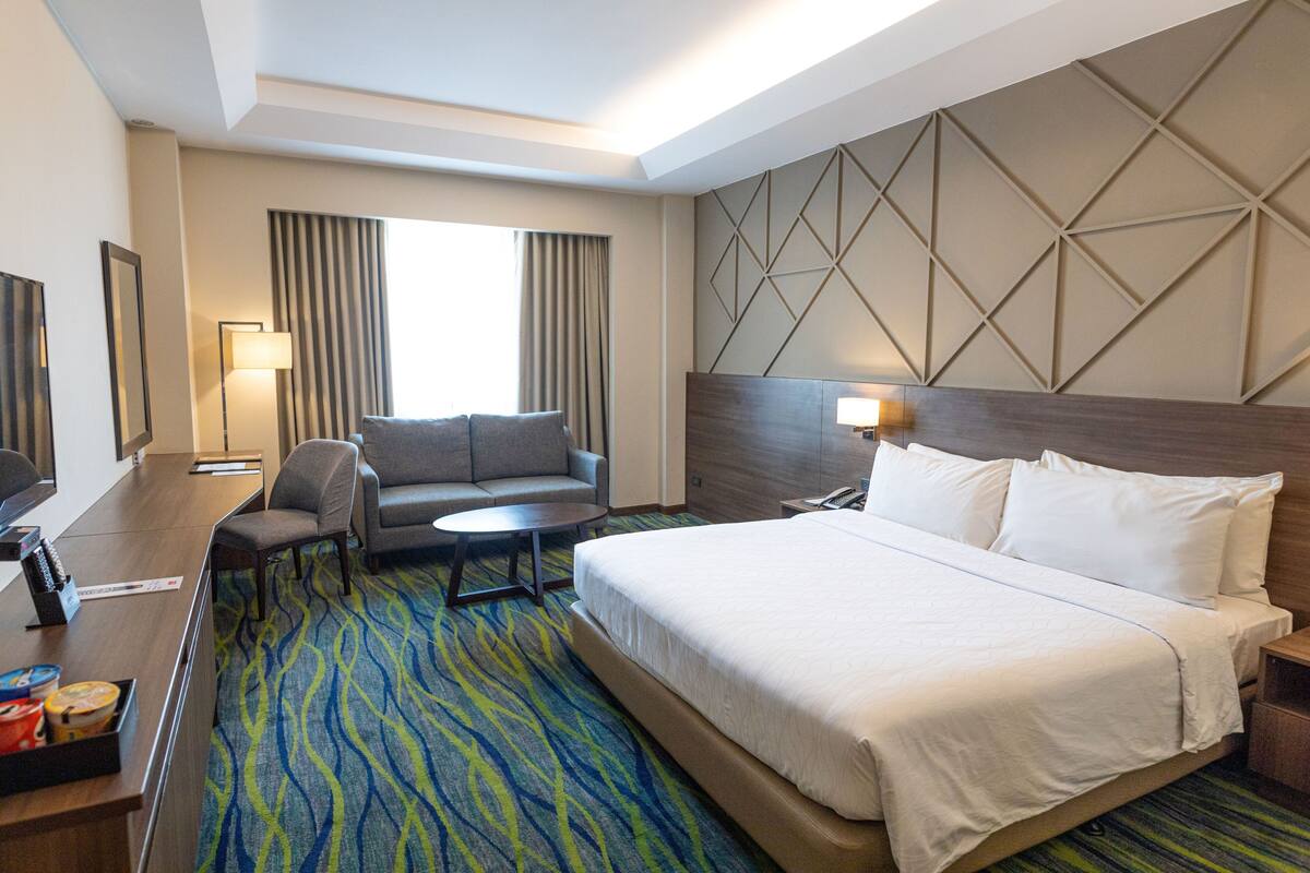 A hotel room with a large bed, a sofa, a desk with a lamp, and a TV.