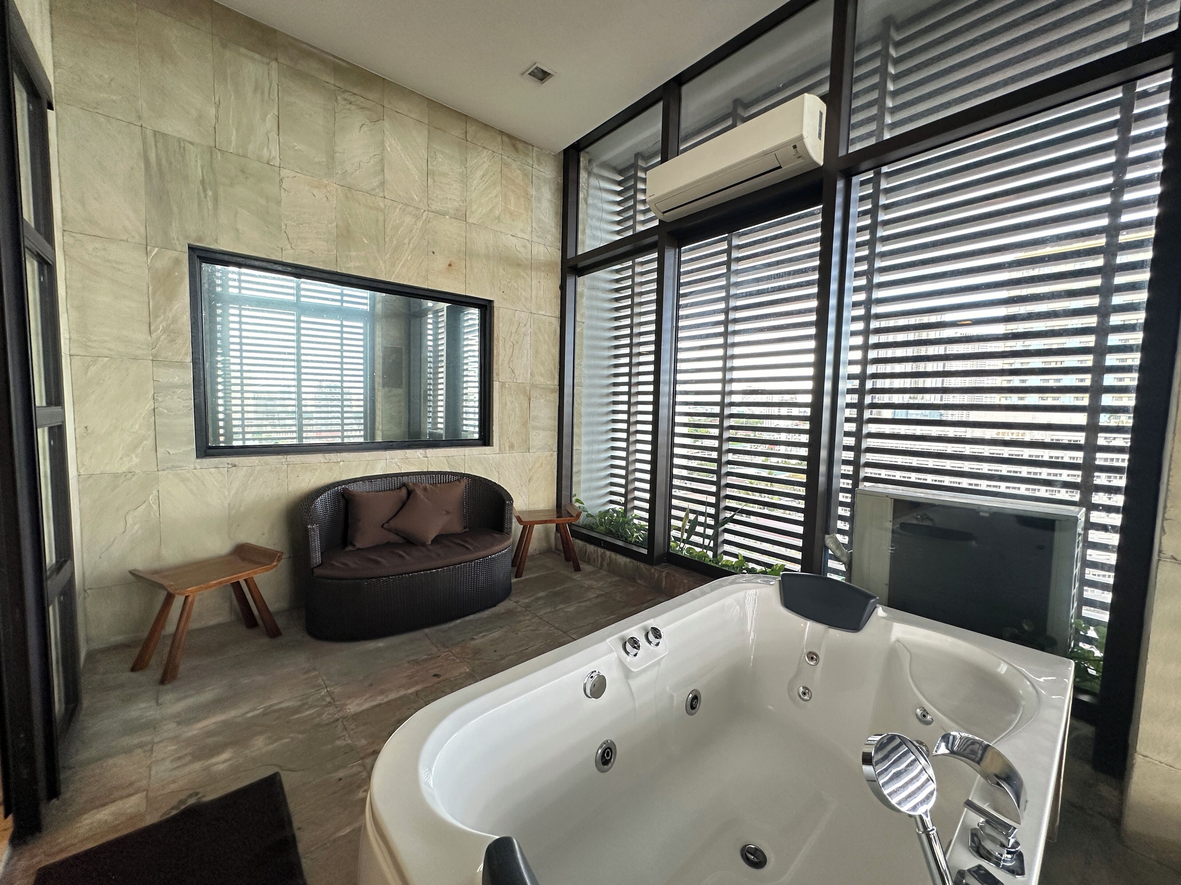 A modern bathroom with a large bathtub, a dark wicker sofa, a small wooden stool, and a large mirror.