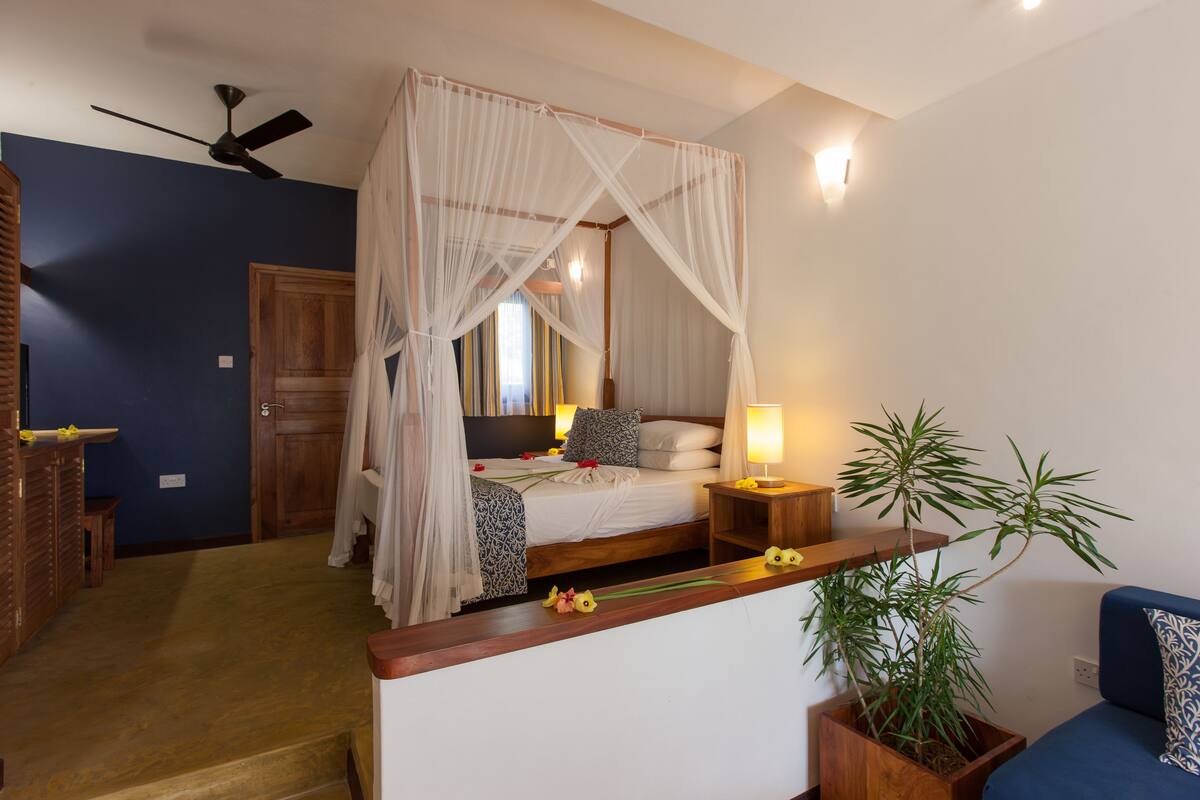 Ein Schlafzimmer mit Himmelbett, Nachttisch mit Lampe, Holzbank, Topfpflanze und blauem Sofa.