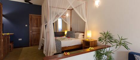 A bedroom with a four-poster bed, a bedside table with a lamp, a wooden bench, a potted plant, and a blue sofa.