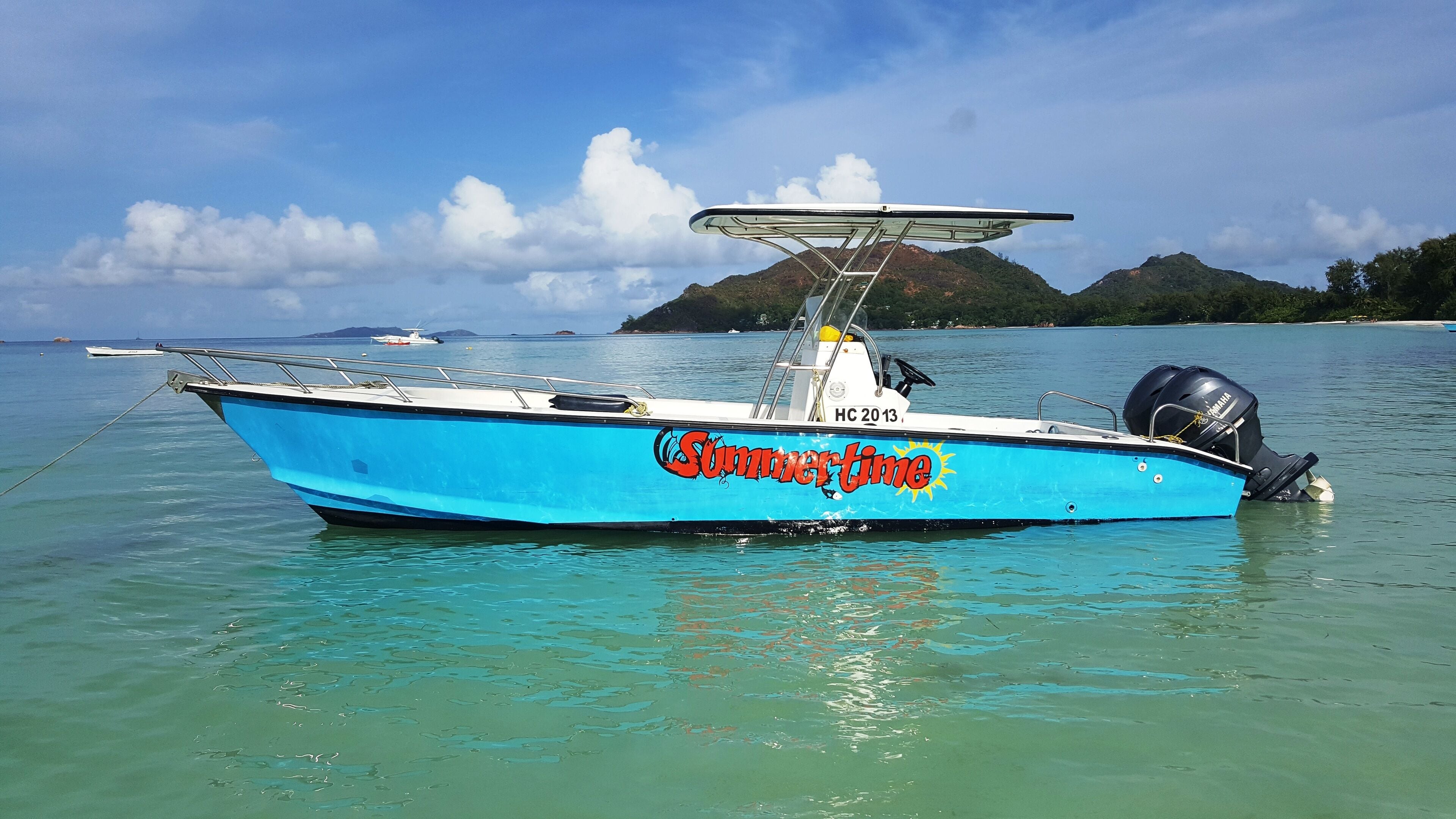 A blue boat with a canopy and a Yamaha outboard motor, anchored in clear turquoise water with a mountainous island in the background.