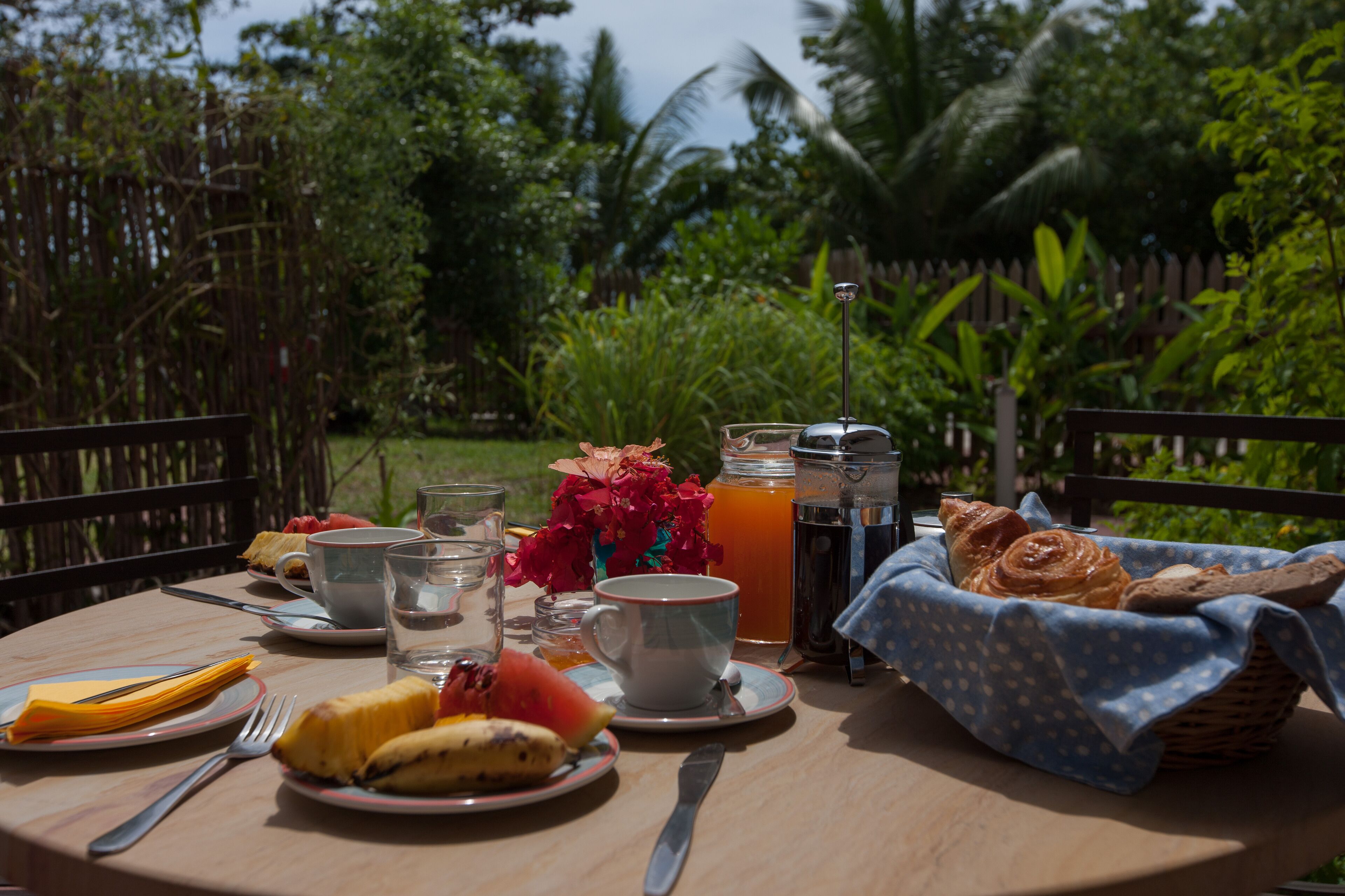 A breakfast table with plates, cups, a basket of bread, and a fruit bowl.
