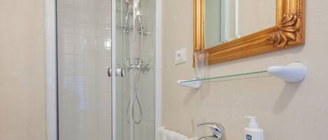 A bathroom with a glass shower enclosure, a white sink with a gold mirror, and a glass shelf above the sink.