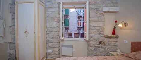 A bedroom with a stone wall, a window with a view of buildings, a wardrobe, a radiator, and a bedside table with a lamp.
