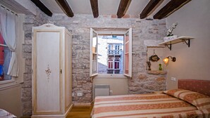 Une chambre à coucher comprenant un lit, une armoire, une fenêtre donnant sur un immeuble et une étagère avec des objets décoratifs.
