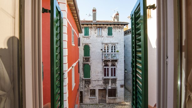 Une ruelle étroite entre deux bâtiments aux volets verts, avec un chemin pavé.