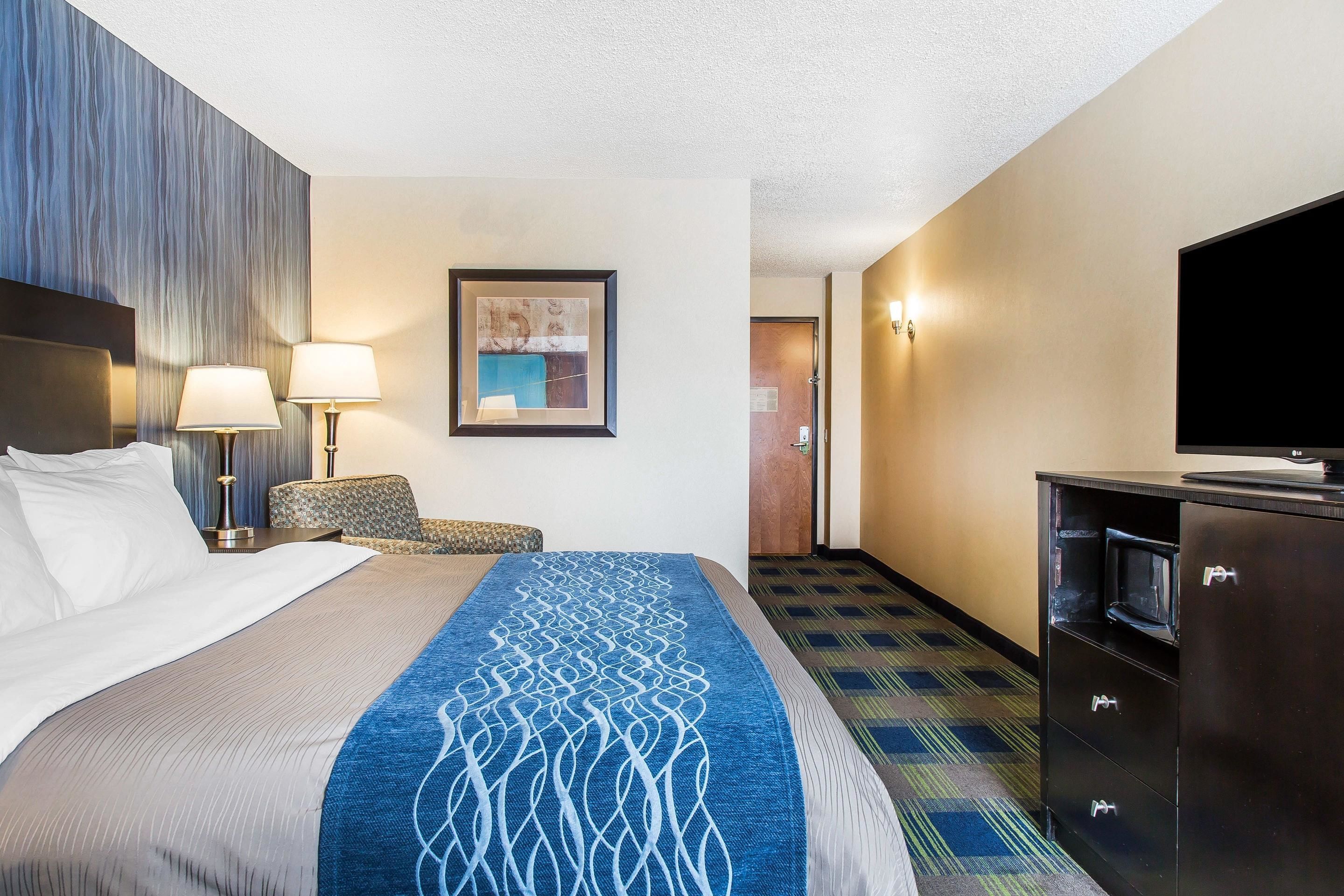 A hotel room with a bed, two bedside lamps, a chair, a television, and a minibar.