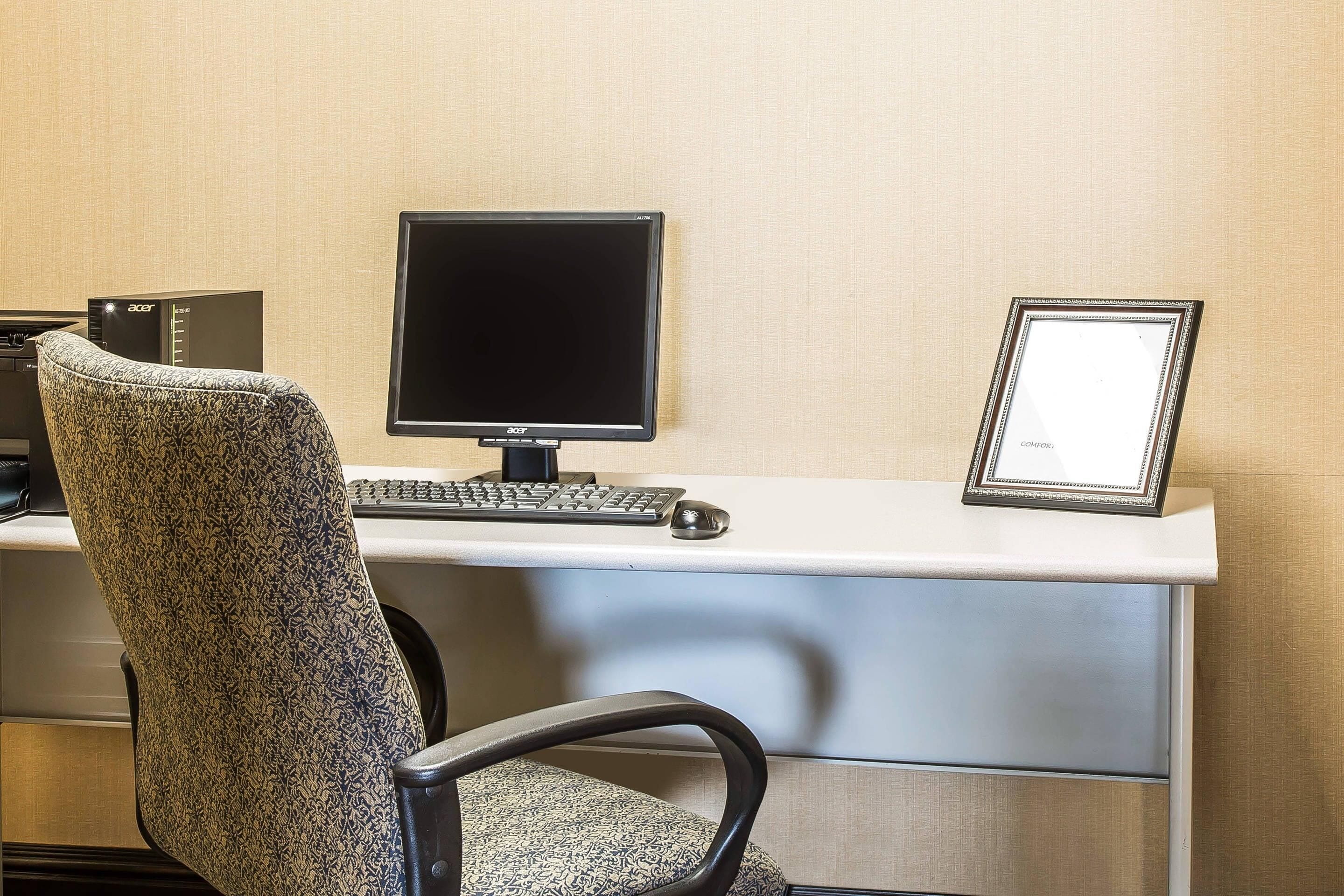 Un espacio de trabajo de hotel con una silla de oficina estampada, un escritorio con monitor de computadora, teclado y ratón, y una fotografía enmarcada sobre el escritorio.