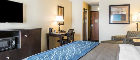 A hotel room with a bed, a desk with a lamp, a microwave, a TV, and a framed picture on the wall.