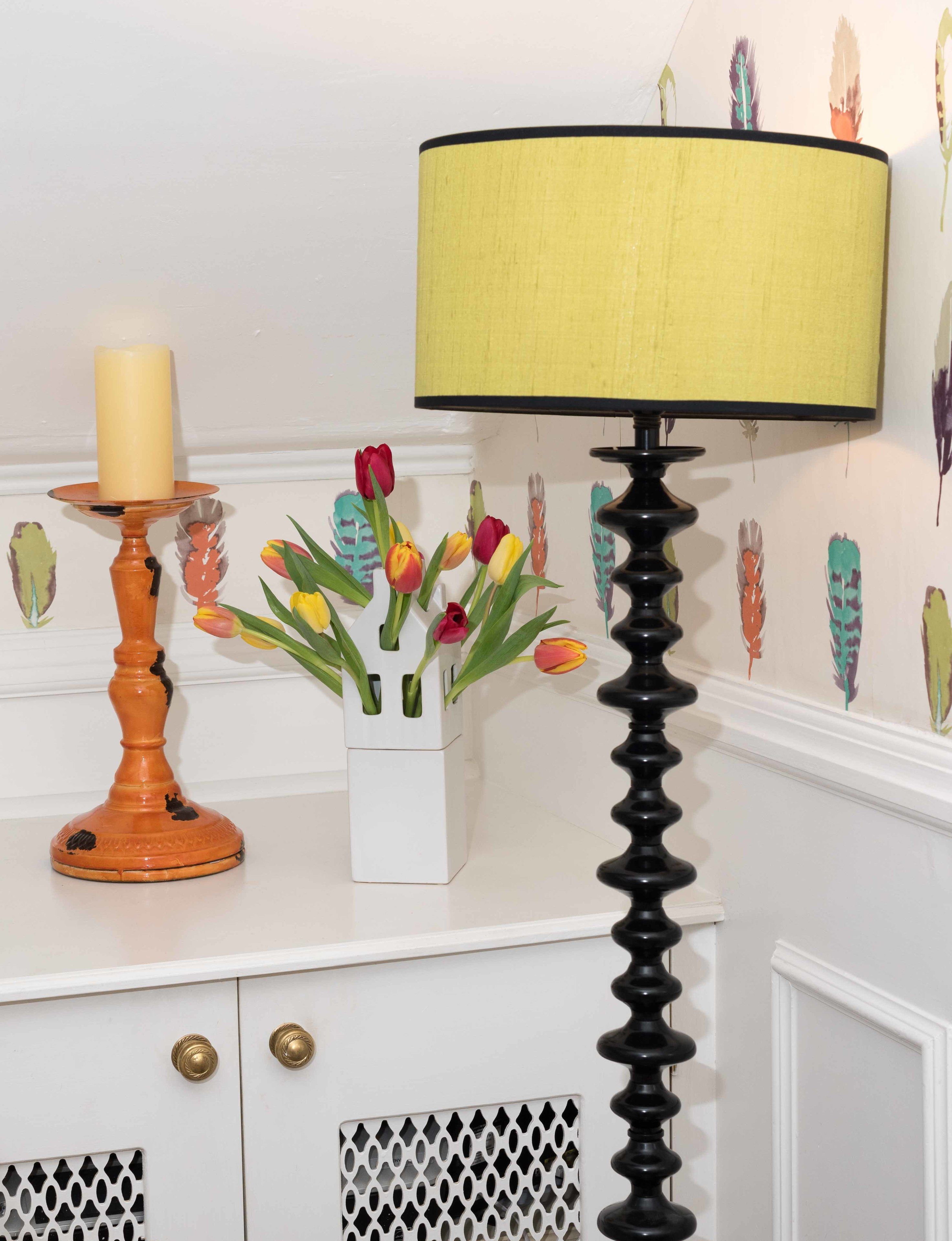 A white cabinet with a decorative vase of tulips, a tall black lamp with a yellow lampshade, and a tall white candle on a wooden stand.