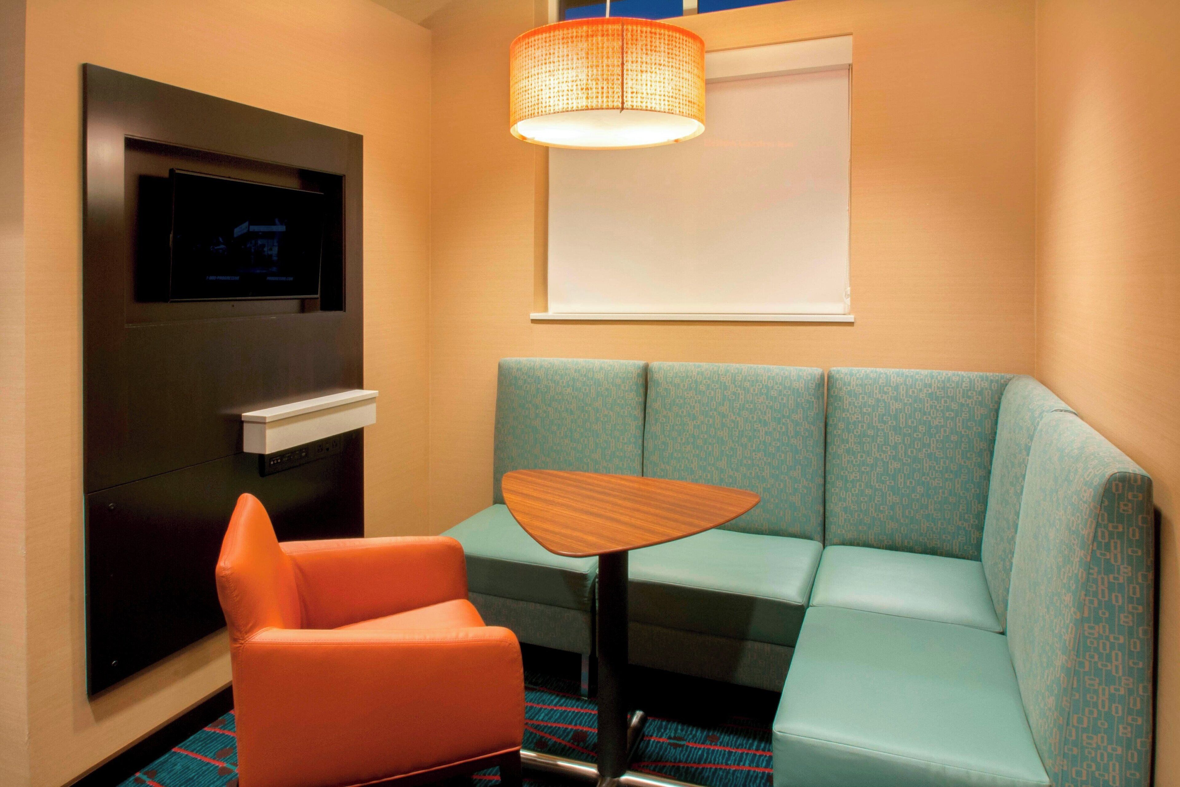 Ein Hotelzimmer mit einem petrolfarbenen Sofa, einem orangefarbenen Sessel, einem kleinen Holztisch, einem flachbildfernseher an der Wand und einem Fenster mit einem weißen Rollo.