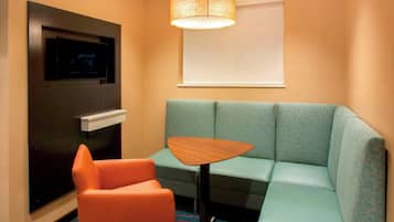 Ein Hotelzimmer mit einem petrolfarbenen Sofa, einem orangefarbenen Sessel, einem kleinen Holztisch, einem flachbildfernseher an der Wand und einem Fenster mit einem weißen Rollo.