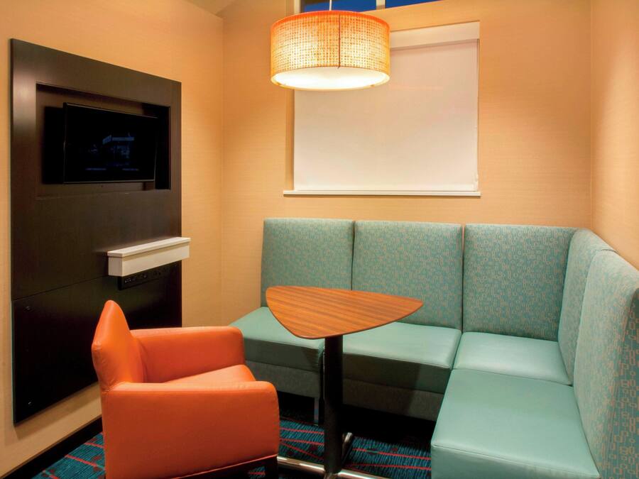 Ein Hotelzimmer mit einem petrolfarbenen Sofa, einem orangefarbenen Sessel, einem kleinen Holztisch, einem flachbildfernseher an der Wand und einem Fenster mit einem weißen Rollo.