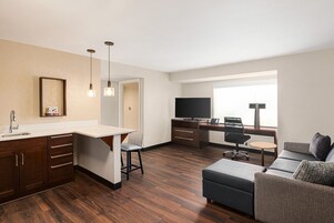 A modern hotel room with a kitchenette, a dining area, a living space with a sofa and a TV, and a large window providing natural light.