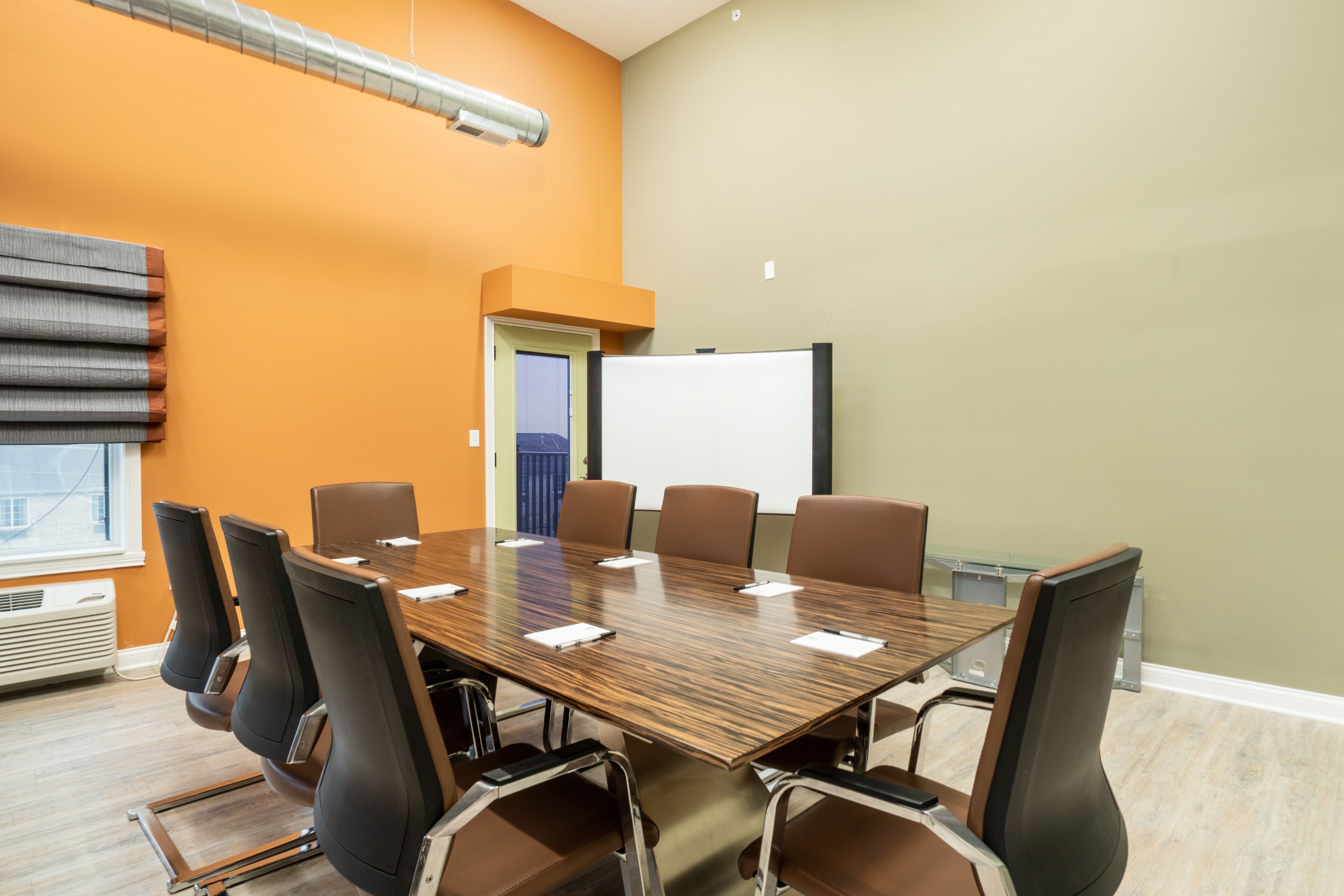 A conference room with a long table, six chairs, a whiteboard, and a window with blinds.
