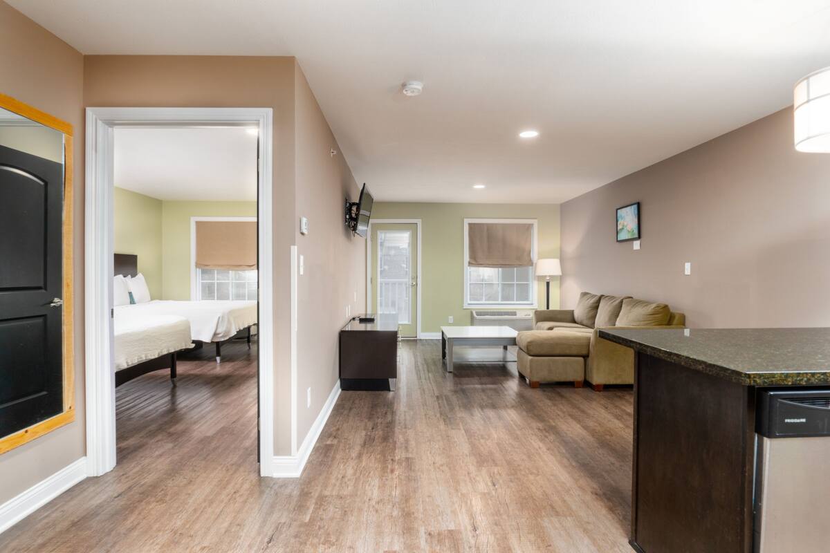 A hotel room with a wooden floor, a bed, a sofa, a dining table, and a kitchen area with a dishwasher.
