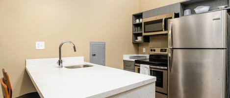 Cocina compacta con encimera blanca, electrodomésticos de acero inoxidable y una silla de madera.