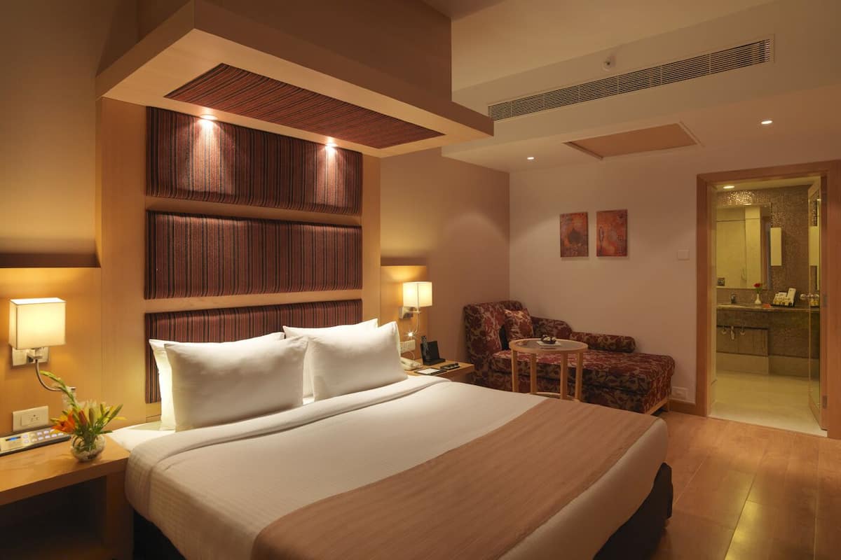 A modern hotel room with a large bed, a seating area with a sofa and armchair, a small table, and a bathroom visible through an open door.