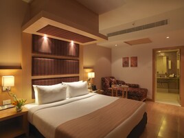 A modern hotel room with a large bed, a seating area with a sofa and armchair, a small table, and a bathroom visible through an open door.
