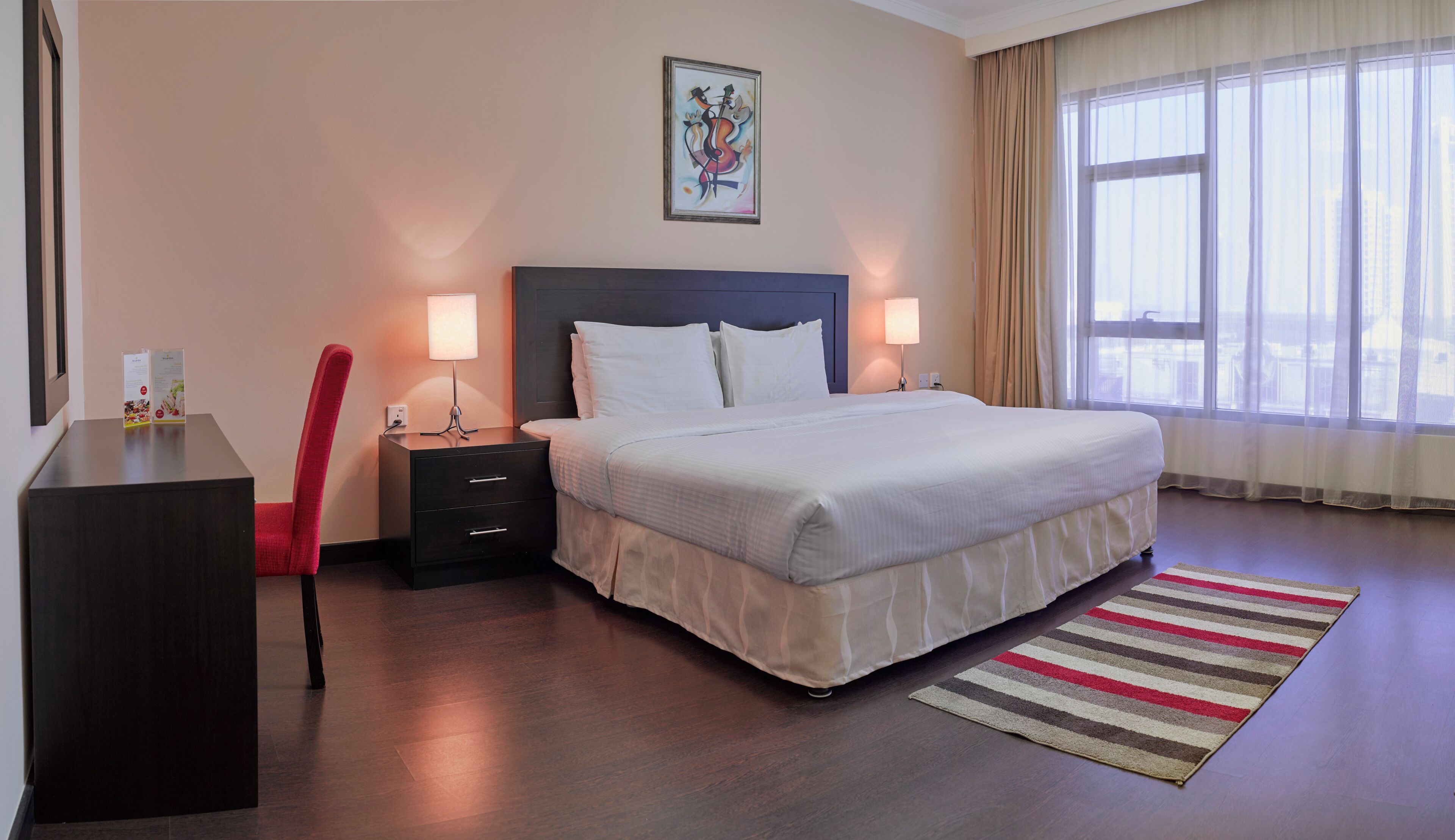 Une chambre d’hôtel comprenant un lit, des tables de chevet, un bureau, une chaise, une fenêtre avec des rideaux et une œuvre d’art accrochée au mur.