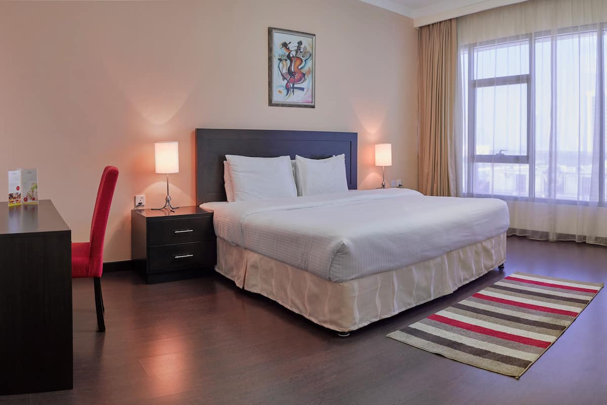 Quarto de hotel com cama, mesas de cabeceira, escrivaninha, cadeira, janela com cortinas e uma obra de arte na parede.