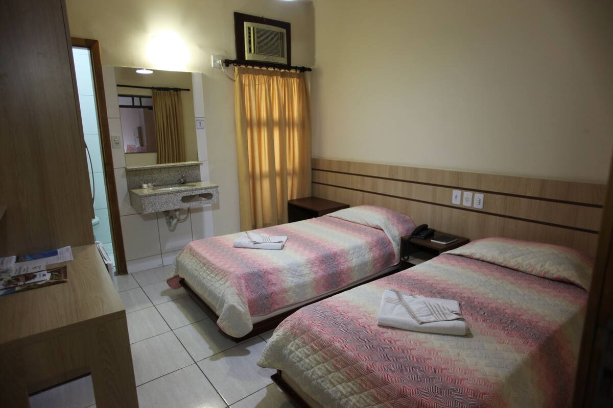 A hotel room with two beds, a wooden headboard, a small bathroom with a mirror, and a window with curtains.