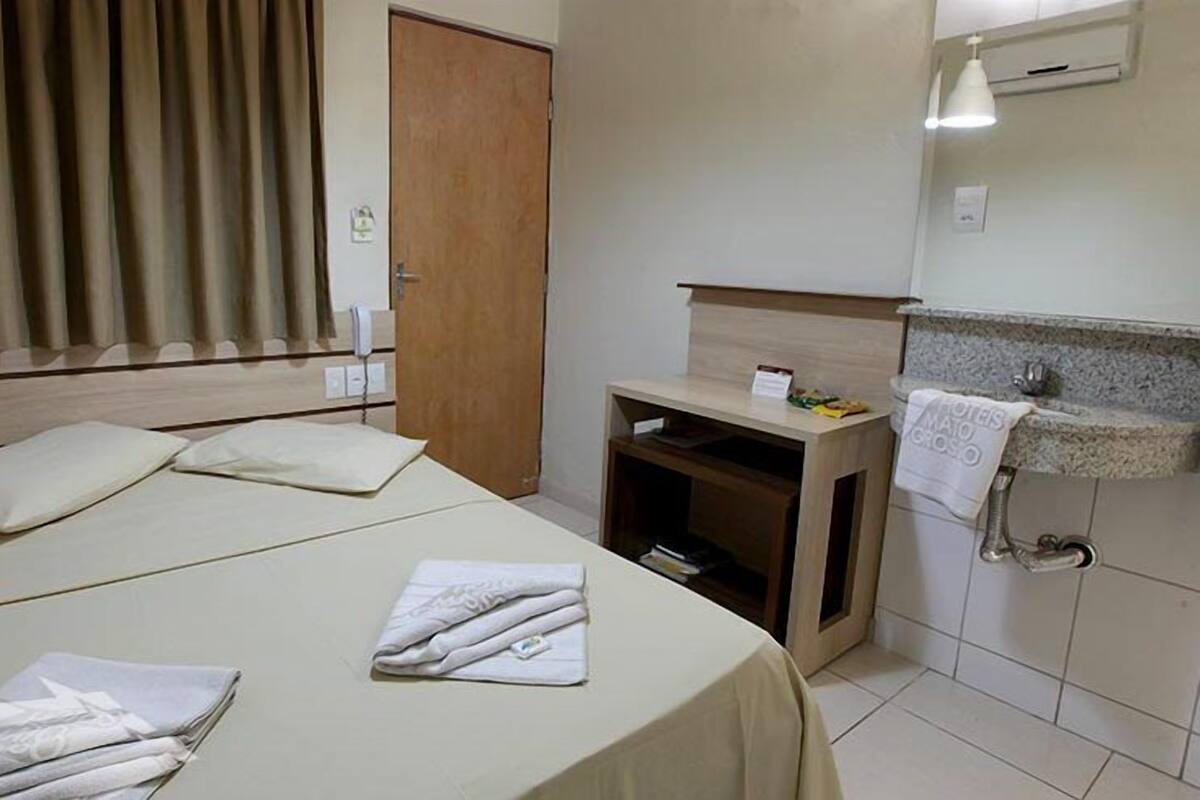 A hotel room with a bed, pillows, a bedside table with a lamp, a small shelf, and a bathroom with a sink and towel rack.