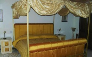 Een hemelbed met een gouden hoofdeinde en voeteinde, geflankeerd door twee nachtkastjes met lampen.