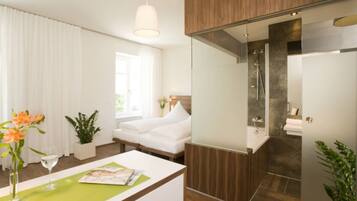 Une chambre d’hôtel moderne avec un lit, un bureau sur lequel se trouve un magazine, un vase avec des fleurs et une vue sur la salle de bain.
