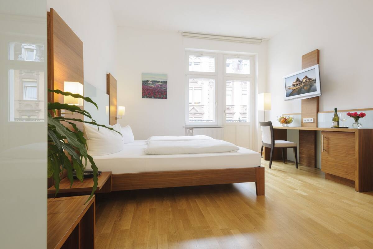 A modern hotel room with a wooden bed, bedside tables, a desk, and a chair. There is a window with a view of buildings and a painting on the wall.