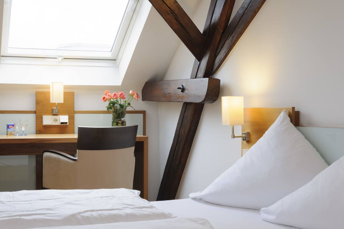 A hotel room with a skylight, a desk with a lamp, a chair, a bed with pillows, and a vase of flowers.