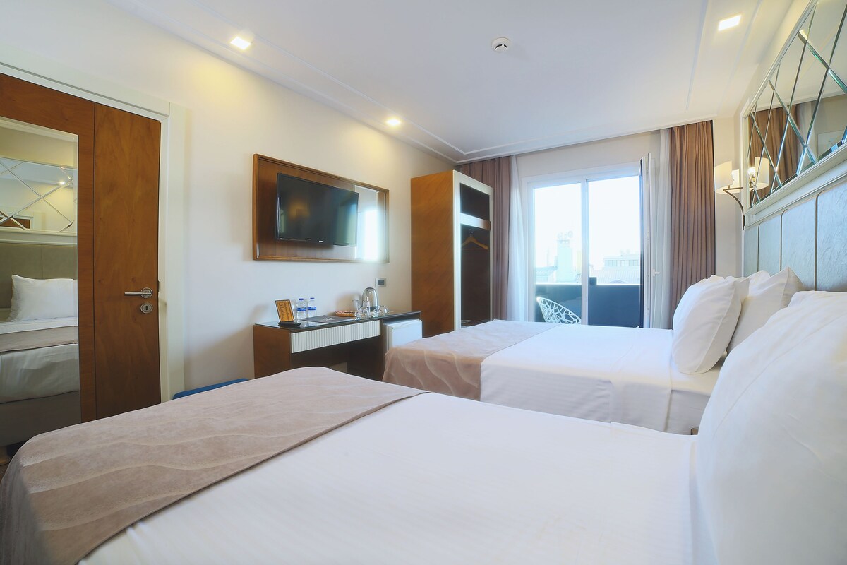 A hotel room with two beds, a desk with a chair, a TV mounted on the wall, and a window with curtains.