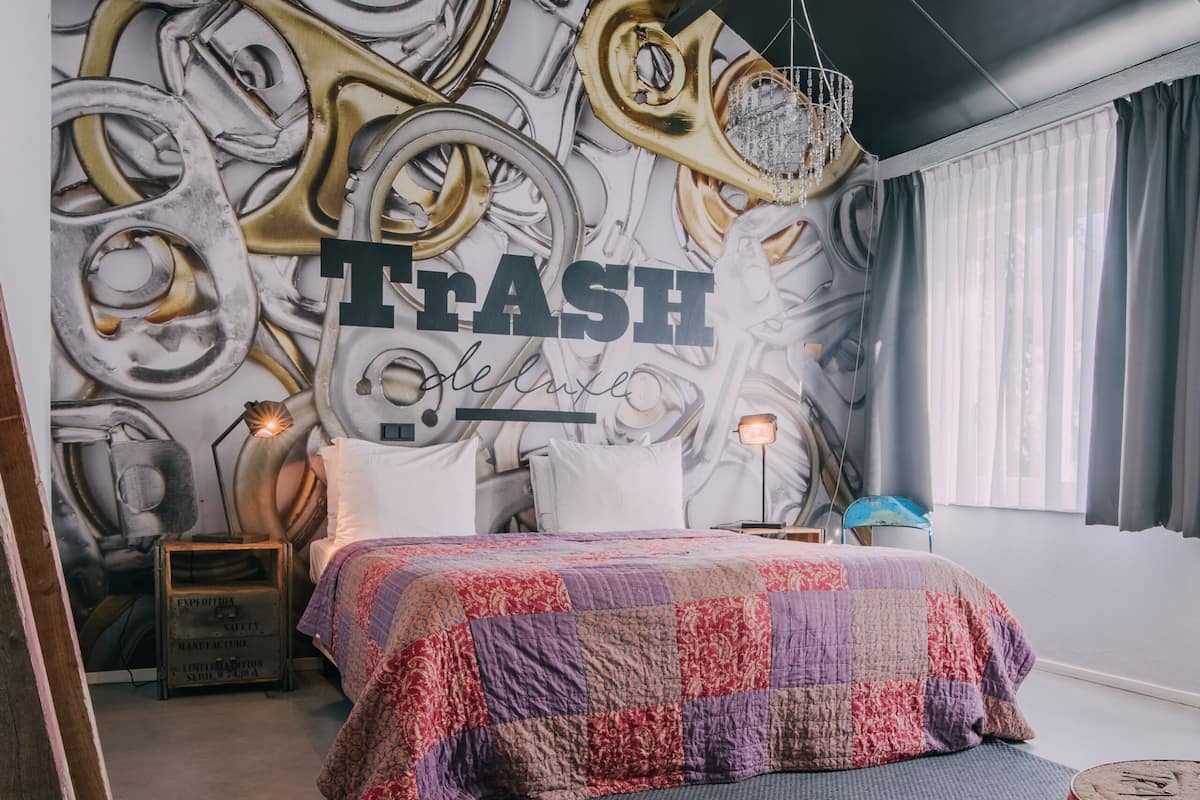 A bedroom with a bed, a wall mural, a hanging light fixture, and a red barrel.